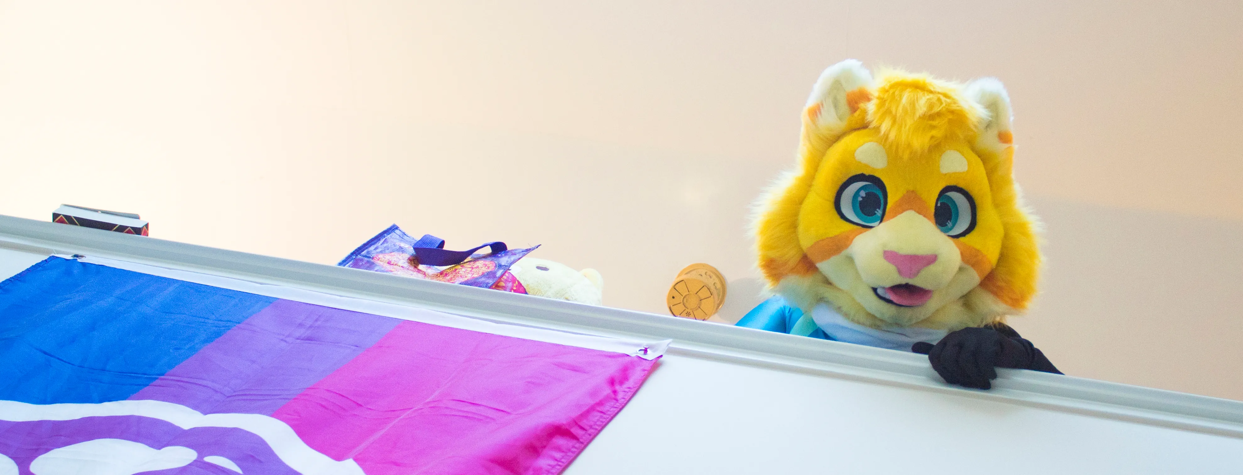 A yellow cat fursuit head peering over the edge of a loft railing, with a bisexual pride flag hanging from the same loft.
